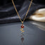 Red CZ Rose Flower Stem Pendant Necklace in gold-tone flat lay on grey mirror surface with dark silk and cream backdrop showing 3D rose bud pendant with faceted red CZ petals and engraved gold stem with leaf details and round bezel CZ accent on double twisted rope chain with mirror reflection below