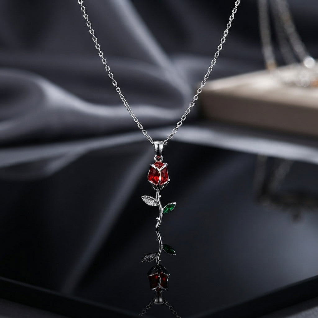 Red CZ Rose Flower Stem Pendant Necklace in silver-tone flat lay on black mirror surface with dark grey silk backdrop showing 3D rose bud pendant with faceted red CZ petals and silver engraved stem with green enamel leaf accent and round bezel CZ on silver cable chain with mirror reflection below