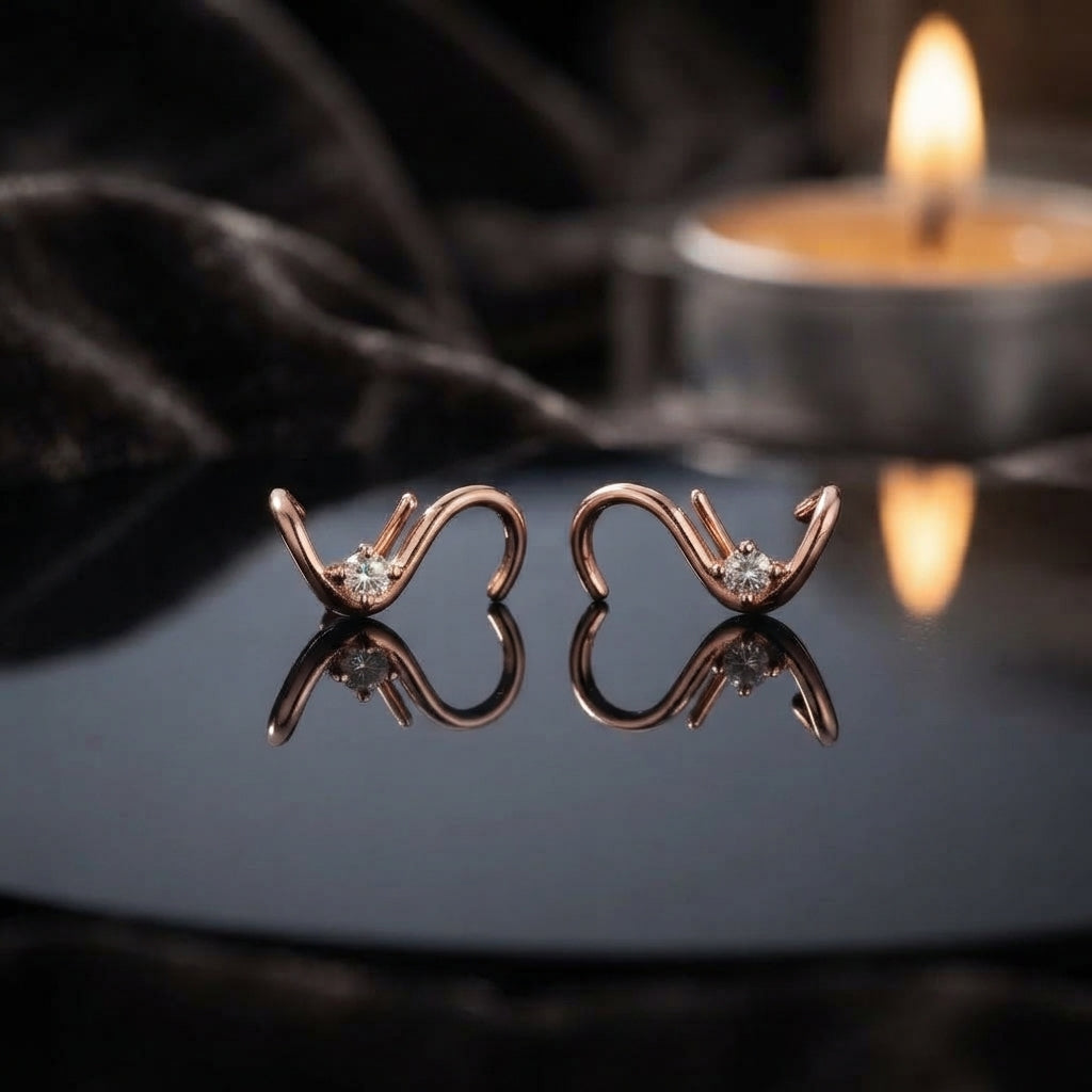 Rose Gold Open Heart CZ Solitaire Stud Earrings — 18K blush rose gold-tone open heart wire frame stud earrings with round brilliant white CZ solitaire in 3-prong setting at heart valley with elegant asymmetric upward curl flourish, displayed on dark grey mirror surface with candle bokeh and dark branch twigs romantic luxury editorial backdrop