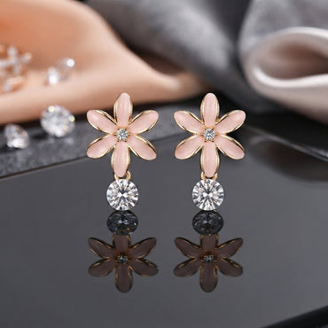 Rose Gold Pink Enamel Flower CZ Drop Stud Earrings pair on black mirror surface with scattered loose CZ stones and grey silk fabric backdrop showing rose gold-tone 5-petal daisy plumeria flower stud with blush pink enamel petals gold outlined edges tiny CZ centre and large round brilliant open-bezel white CZ drop with mirror reflections below