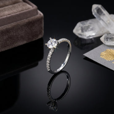 Silver-Tone 4-Prong Round CZ Solitaire Full Pavé Eternity Band Engagement Ring on black reflective surface with taupe velvet ring box and white quartz crystal clusters backdrop