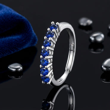 Silver-Tone 7-Stone Round Blue Sapphire CZ Half Eternity Adjustable Band Ring on black reflective surface with navy blue velvet pouch and scattered black pebbles backdrop