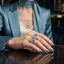 Silver Aqua Blue and Sapphire CZ Peacock Feather Open Adjustable Statement Ring worn on index finger of female model in steel blue satin blazer and cream camisole with blue silk neck scarf in dark luxury boutique backdrop with gold shelving showing oval aqua blue Paraiba teal CZ apex sapphire blue CZ mid-accent and full micro-pave white CZ peacock feather design in rhodium silver