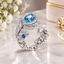 Silver Aqua Blue and Sapphire CZ Peacock Feather Open Adjustable Statement Ring standing on white Carrara marble surface with white gypsophila flowers and cream gold gift box backdrop showing full 360 degree design with oval aqua blue Paraiba teal CZ solitaire apex round sapphire blue CZ mid-stem accent and fully micro-pave white CZ set peacock feather head and open sweeping pave band arms in rhodium silver