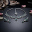 Silver Baguette Fan Scallop CZ Necklace Earrings Tikka Bridal Set in Emerald Green — flat-lay of rhodium-plated 3-piece bridal set: emerald green baguette CZ scallop necklace, chandelier earrings and maang tikka on dark mirror surface with rose petals editorial backdrop