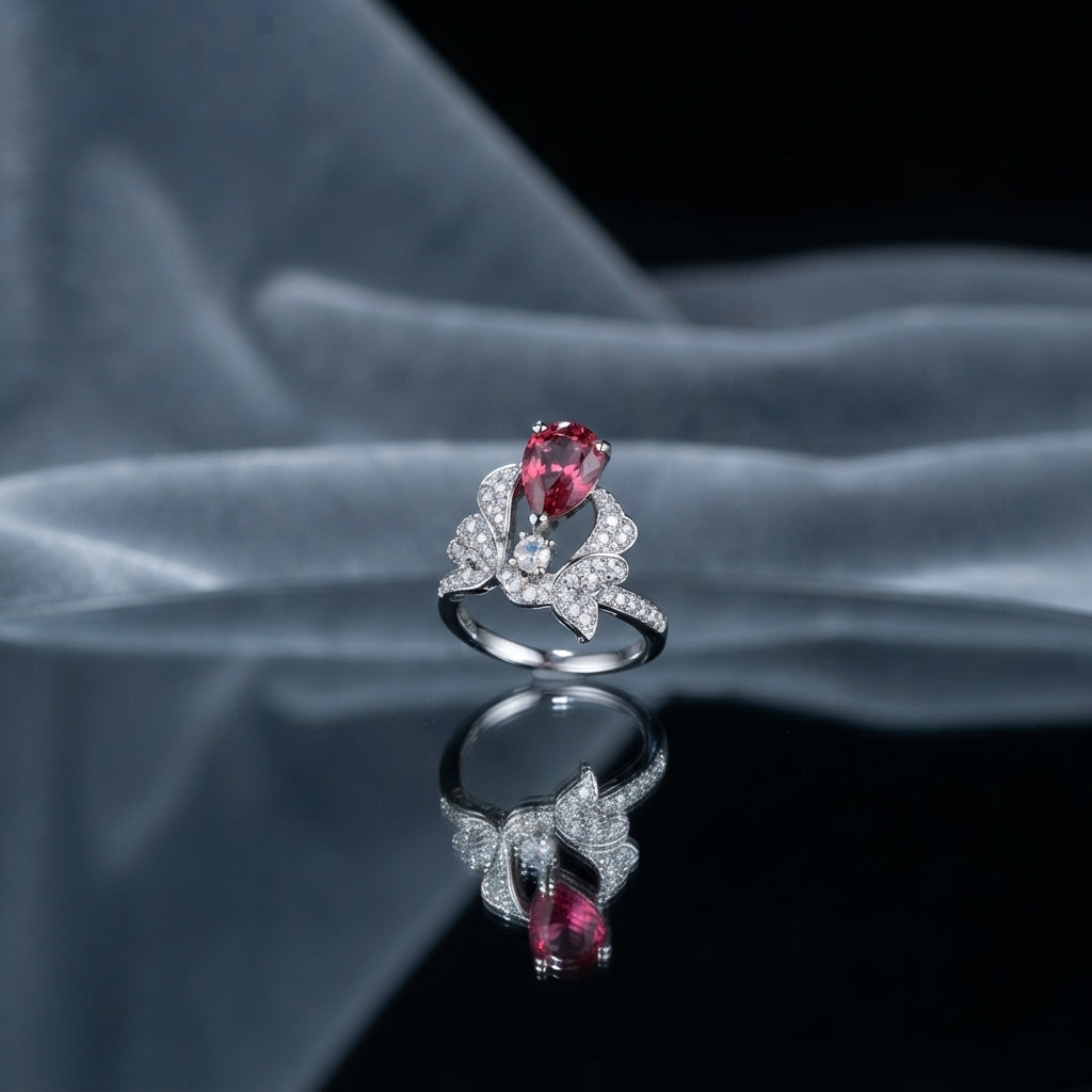 Silver Butterfly Ruby Red Pear CZ Pavé Cocktail Ring — angled 3/4 view: rhodium butterfly ring with ruby-red pear center, full CZ pavé wings, CZ shoulder band, on grey silk fabric with mirror reflection and soft architectural editorial backdrop