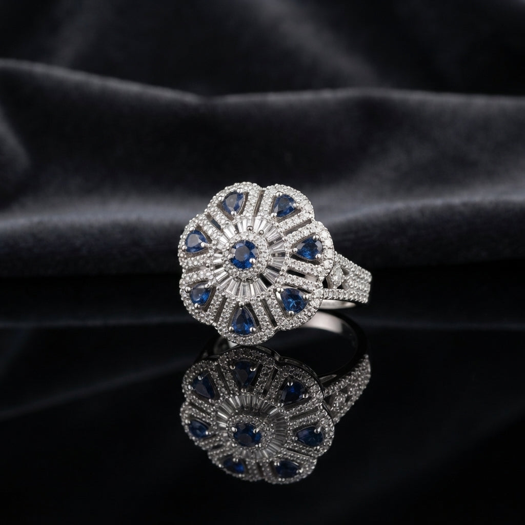 Silver CZ Art Deco Sunburst Flower Pear Petal Pavé Cocktail Ring in Blue Sapphire — deep navy sapphire blue CZ pear petals and round center on wide white pavé split shank, luxury product shot on black mirror surface with dark charcoal silk backdrop
