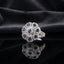Silver CZ Art Deco Sunburst Flower Pear Petal Pavé Cocktail Ring in Blue Sapphire — deep navy sapphire blue CZ pear petals and round center on wide white pavé split shank, luxury product shot on black mirror surface with dark charcoal silk backdrop