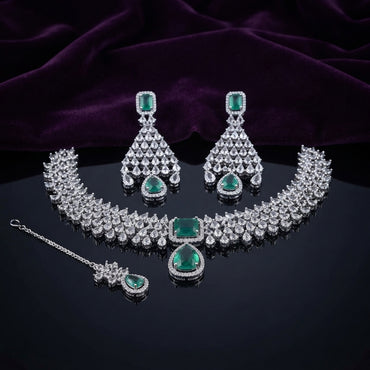 Silver CZ Cluster Choker Earrings and Maang Tikka Bridal Set in Emerald Green on dark mirror surface with purple velvet backdrop showing rhodium silver wide white CZ cluster choker with emerald green CZ square and pear pendant halo matching fan-drop earrings and maang tikka
