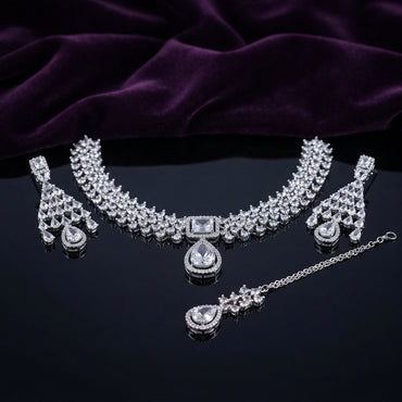 Silver CZ Cluster Choker Earrings and Maang Tikka Bridal Set in Diamond White on dark mirror surface with purple velvet backdrop showing rhodium silver wide mixed-cut white CZ cluster choker with white CZ square and pear pendant halo fan-drop earrings and maang tikka