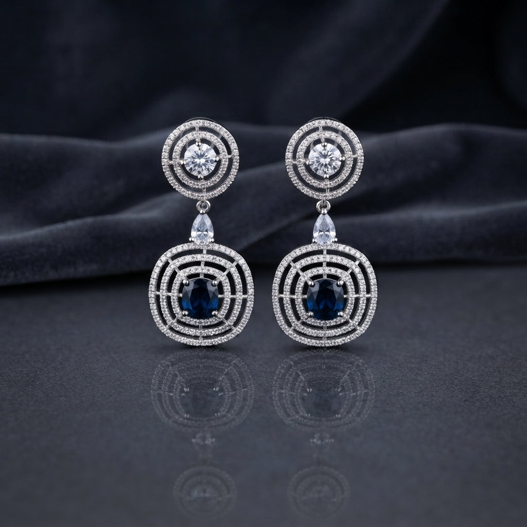 Silver CZ Concentric Orbit Double Disc and Pear Connector Drop Earrings in Aqua Blue on dark mirror surface with charcoal satin backdrop showing rhodium silver pair with concentric cushion orbit stud white pear connector and large orbit disc pendant with oval aqua blue teal cabochon CZ centre