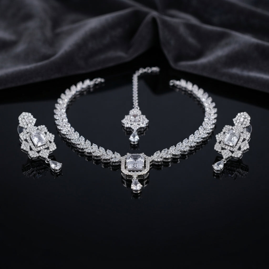 Silver CZ Diamond-Illusion Bridal 3-Piece Jewelry Set on black mirror surface with dark grey silk backdrop showing V-shape CZ leaf chain necklace with large emerald-cut centre halo pendant and teardrop drop flanked by matching drop earrings with emerald-cut CZ halo top and teardrop drop and small maang tikka with CZ halo and teardrop drop with mirror reflection below