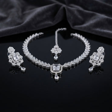 Silver CZ Diamond-Illusion Bridal 3-Piece Jewelry Set on black mirror surface with dark grey silk backdrop showing V-shape CZ leaf chain necklace with large emerald-cut centre halo pendant and teardrop drop flanked by matching drop earrings with emerald-cut CZ halo top and teardrop drop and small maang tikka with CZ halo and teardrop drop with mirror reflection below
