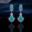 Silver CZ Double Teardrop Cabochon Lotus Fan Chandelier Earrings in Emerald Green pair on dark mirror surface with grey silk backdrop showing rhodium-plated small emerald green pear cabochon stud with white CZ pave halo infinity connector and large emerald green pear cabochon drop with white CZ pave halo and marquise CZ lotus fan crown with mirror reflection below