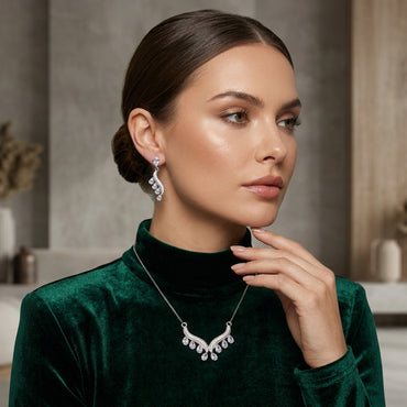 Silver White CZ Feather Wing V-Bib Necklace and Drop Earrings Set worn by model in emerald green velvet turtleneck showing all-white diamond illusion CZ feather wing V-bib necklace and matching feather wing drop earrings in warm grey editorial setting