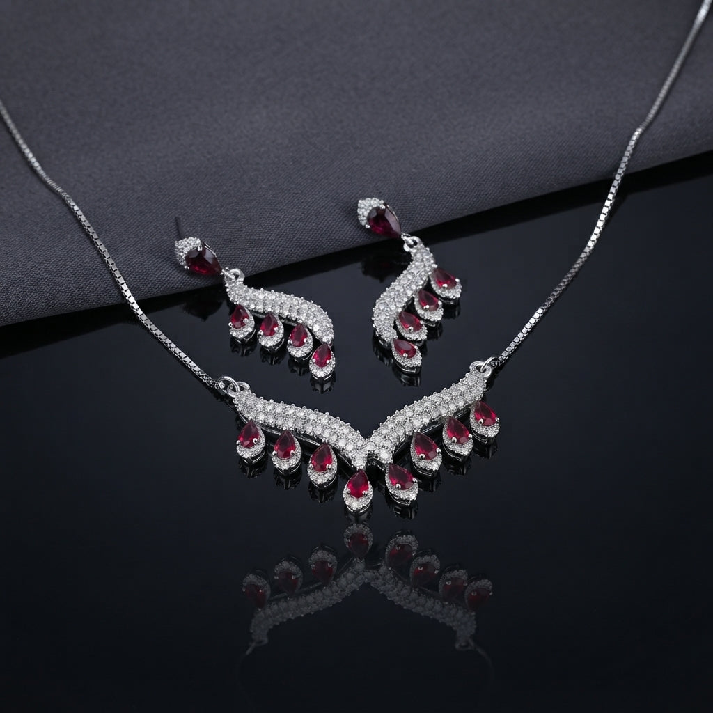 Silver Sapphire Blue CZ Feather Wing V-Bib Necklace and Drop Earrings Set flat lay on dark grey draped fabric showing navy sapphire blue CZ graduating pear drops with white CZ micro-pavé feather wing body necklace and matching earrings with mirror reflection