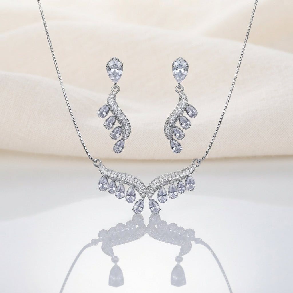 Silver White CZ Feather Wing V-Bib Necklace and Drop Earrings Set flat lay on ivory cream satin backdrop showing all-white diamond illusion CZ V-bib necklace with graduating pear CZ drops and matching feather wing drop earrings with reflection