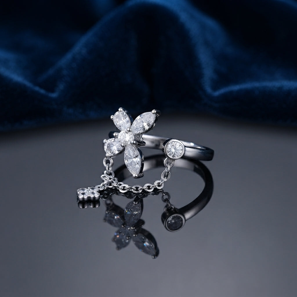 Silver CZ Marquise Flower and Bezel Round Chain Drop Open Adjustable Ring standing on dark grey mirror surface with deep navy teal satin velvet backdrop showing 4-petal marquise-cut white CZ flower top with round CZ centre connected by fine cable chain with small 4-petal CZ flower charm pendant to flat bezel-set round CZ opposite open arm in rhodium silver with dark oxidized shank and full mirror reflection below