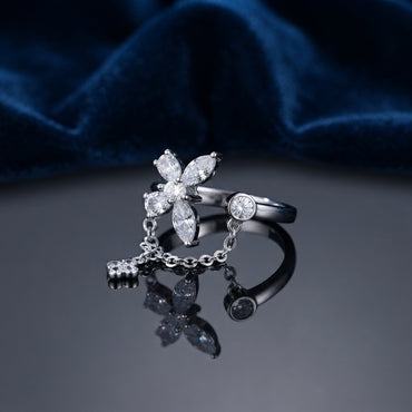 Silver CZ Marquise Flower and Bezel Round Chain Drop Open Adjustable Ring standing on dark grey mirror surface with deep navy teal satin velvet backdrop showing 4-petal marquise-cut white CZ flower top with round CZ centre connected by fine cable chain with small 4-petal CZ flower charm pendant to flat bezel-set round CZ opposite open arm in rhodium silver with dark oxidized shank and full mirror reflection below