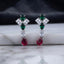 Silver CZ Pinwheel and Pear Ruby Drop Earrings in Rose Pink and Ruby colorway on dark mirror surface with charcoal satin backdrop showing rhodium silver princess CZ pinwheel cluster with rose pink oval and marquise accents baguette white CZ connector and vivid ruby red pear CZ drop