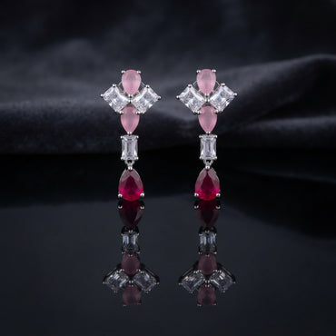 Silver CZ Pinwheel and Pear Ruby Drop Earrings in Emerald and Ruby colorway on white marble surface with dark satin backdrop showing rhodium silver princess CZ pinwheel cluster with emerald green oval and marquise accents baguette white CZ connector and vivid ruby red pear CZ drop