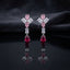 Silver CZ Pinwheel and Pear Ruby Drop Earrings in Emerald and Ruby colorway on white marble surface with dark satin backdrop showing rhodium silver princess CZ pinwheel cluster with emerald green oval and marquise accents baguette white CZ connector and vivid ruby red pear CZ drop