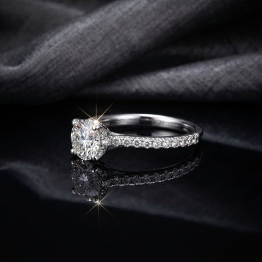 Silver CZ Solitaire Pavé Shoulder Engagement Ring side profile view on black mirror surface with dark grey fabric backdrop showing round brilliant CZ solitaire in 4-claw rhodium prong setting on slim tapered shank with single-row micro-pave CZ shoulder stones with sparkle starburst effect and mirror reflection below