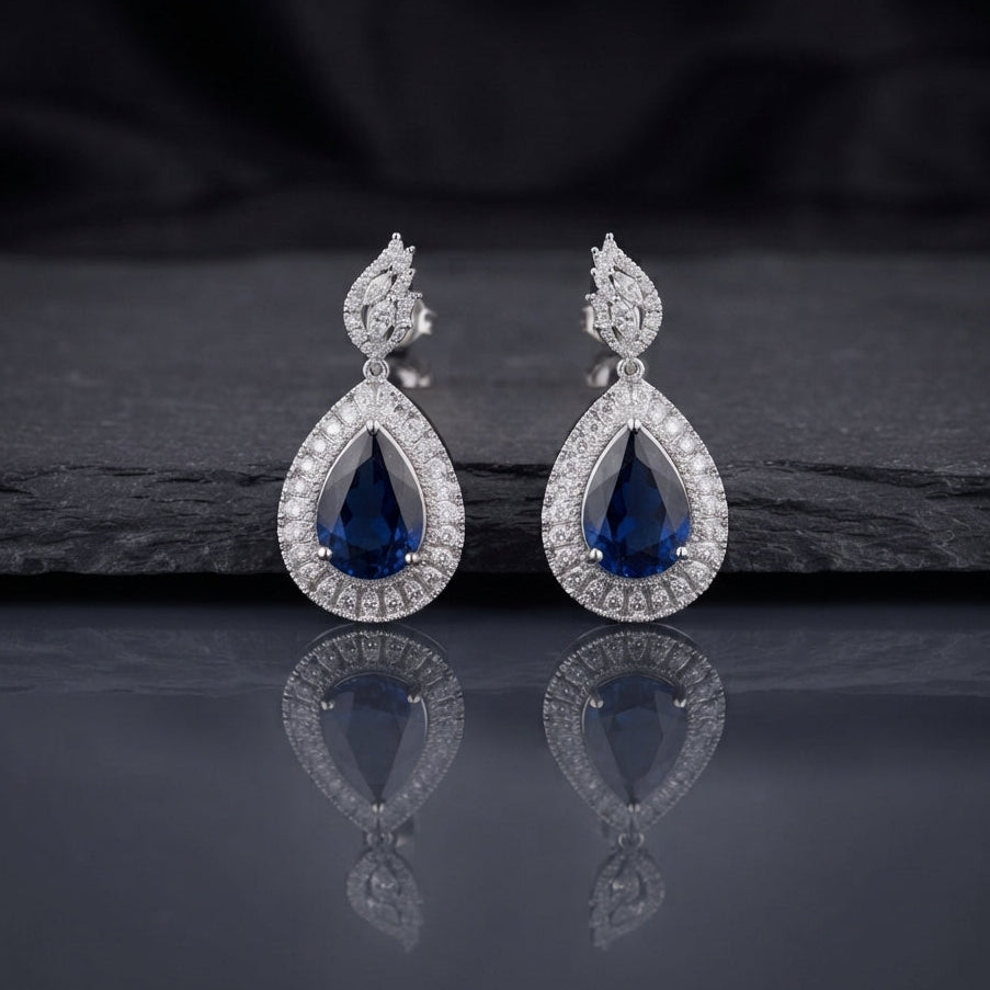 Silver AD Teardrop Halo Drop Earrings in Sapphire Blue pair on dark slate mirror surface showing rhodium-plated marquise AD leaf cluster stud top and large open pear teardrop frame with round AD pave halo border and deep sapphire blue faceted pear AD centre stone with mirror reflection below
