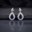 Silver AD Teardrop Halo Drop Earrings in Sapphire Blue pair on dark slate mirror surface showing rhodium-plated marquise AD leaf cluster stud top and large open pear teardrop frame with round AD pave halo border and deep sapphire blue faceted pear AD centre stone with mirror reflection below