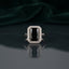 Silver Emerald Cut CZ Rectangular Halo Pave Band Solitaire Ring in Emerald Green variant on dark mirror surface with charcoal satin backdrop showing rich deep emerald green emerald-cut CZ solitaire in 4-claw setting with full rectangular white CZ halo and continuous micro-pave white CZ band in rhodium silver with mirror reflection below