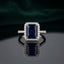 Silver Emerald Cut CZ Rectangular Halo Pave Band Solitaire Ring in Jet Black Onyx variant on dark mirror surface with dark teal velvet backdrop showing bold opaque jet black emerald-cut CZ solitaire in 4-claw setting with full rectangular white CZ halo and continuous micro-pave white CZ band in rhodium silver with mirror reflection below