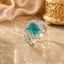 silver-Tone Emerald-Cut Green CZ Halo & Cluster Shoulder Cocktail Ring on cream marble surface with champagne satin fabric and gold Mughal decorative urn backdrop