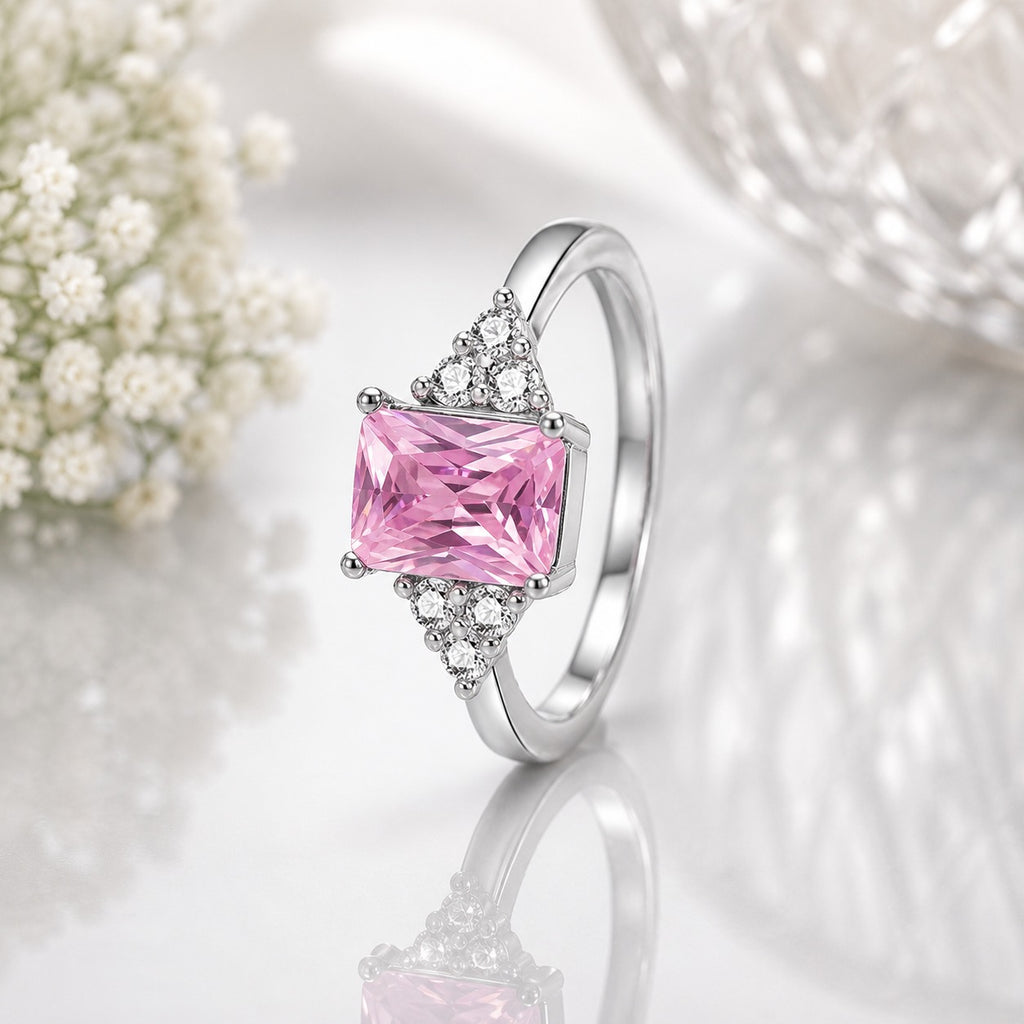 Silver-Tone Emerald-Cut Pink CZ Solitaire Ring with Triangle White CZ Accents on white reflective surface with baby's breath flowers backdrop