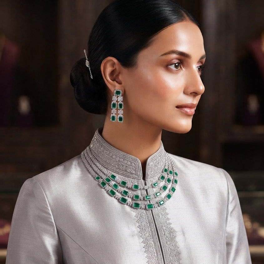 Silver Emerald Green CZ 3-Layer Emerald-Cut Tennis Necklace and Earrings Luxury Bridal Set worn by South Asian model in silver grey silk kurta with embroidered neckline in luxury jewelry store setting showing triple-strand layered collar necklace and long 3-stone drop earrings with emerald green CZ stones