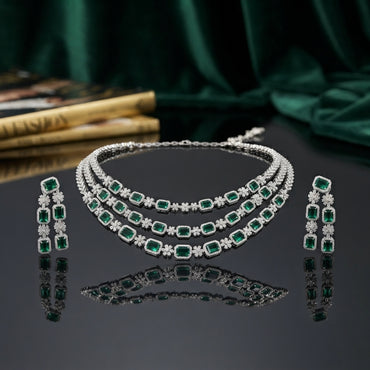 Silver Emerald Green CZ 3-Layer Emerald-Cut Tennis Necklace and Earrings Luxury Bridal Set on dark grey mirror surface with emerald green silk backdrop and fashion books showing triple-strand layered collar necklace with emerald-cut green CZ square halo stations and white CZ daisy rosette spacers flanked by matching 3-stone vertical drop earrings with mirror reflection below
