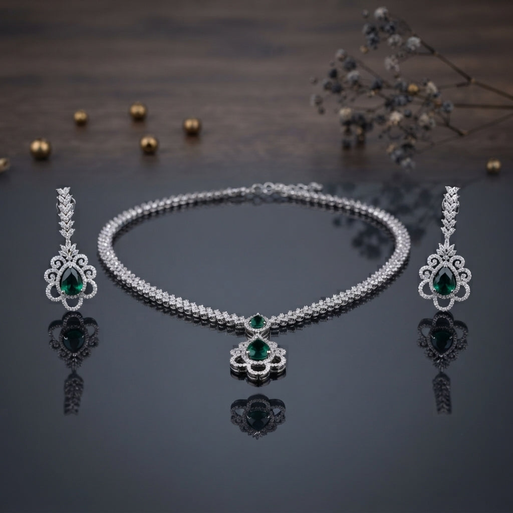 Silver Emerald Green CZ Floral Scroll Pendant Necklace and Earrings 2-Piece Set on dark grey mirror surface with dark wood backdrop gold pearls and dried botanicals showing double-row CZ tennis necklace with open baroque scroll floral pendant and large emerald green pear CZ centre flanked by matching long chandelier drop earrings with CZ leaf spray top and emerald green pear CZ centre with mirror reflections below