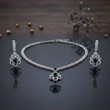 Silver Emerald Green CZ Floral Scroll Pendant Necklace and Earrings 2-Piece Set on dark grey mirror surface with dark wood backdrop gold pearls and dried botanicals showing double-row CZ tennis necklace with open baroque scroll floral pendant and large emerald green pear CZ centre flanked by matching long chandelier drop earrings with CZ leaf spray top and emerald green pear CZ centre with mirror reflections below