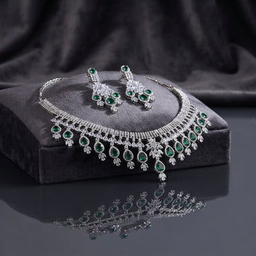 Silver Emerald Green CZ Fringe Tassel Bridal Necklace and Earrings 2-Piece Luxury Set on dark charcoal stone display block with dark grey silk backdrop showing grand V-fringe statement necklace with double-row baguette CZ top bar and graduated emerald green pear CZ tassel drops with matching chandelier drop earrings featuring emerald green CZ cluster and teardrop drops with mirror reflection below