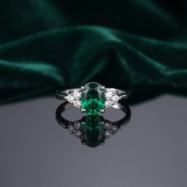 Silver Emerald Green Oval CZ and White CZ Cluster Shoulder Cocktail Ring flat lay on grey mirror surface with dark emerald green velvet backdrop showing rhodium silver ring with large oval brilliant-cut deep emerald green CZ solitaire in 4-claw prong setting and 3-stone white CZ triangular cluster shoulders on each side with mirror reflection below