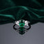 Silver Emerald Green Oval CZ and White CZ Cluster Shoulder Cocktail Ring flat lay on grey mirror surface with dark emerald green velvet backdrop showing rhodium silver ring with large oval brilliant-cut deep emerald green CZ solitaire in 4-claw prong setting and 3-stone white CZ triangular cluster shoulders on each side with mirror reflection below