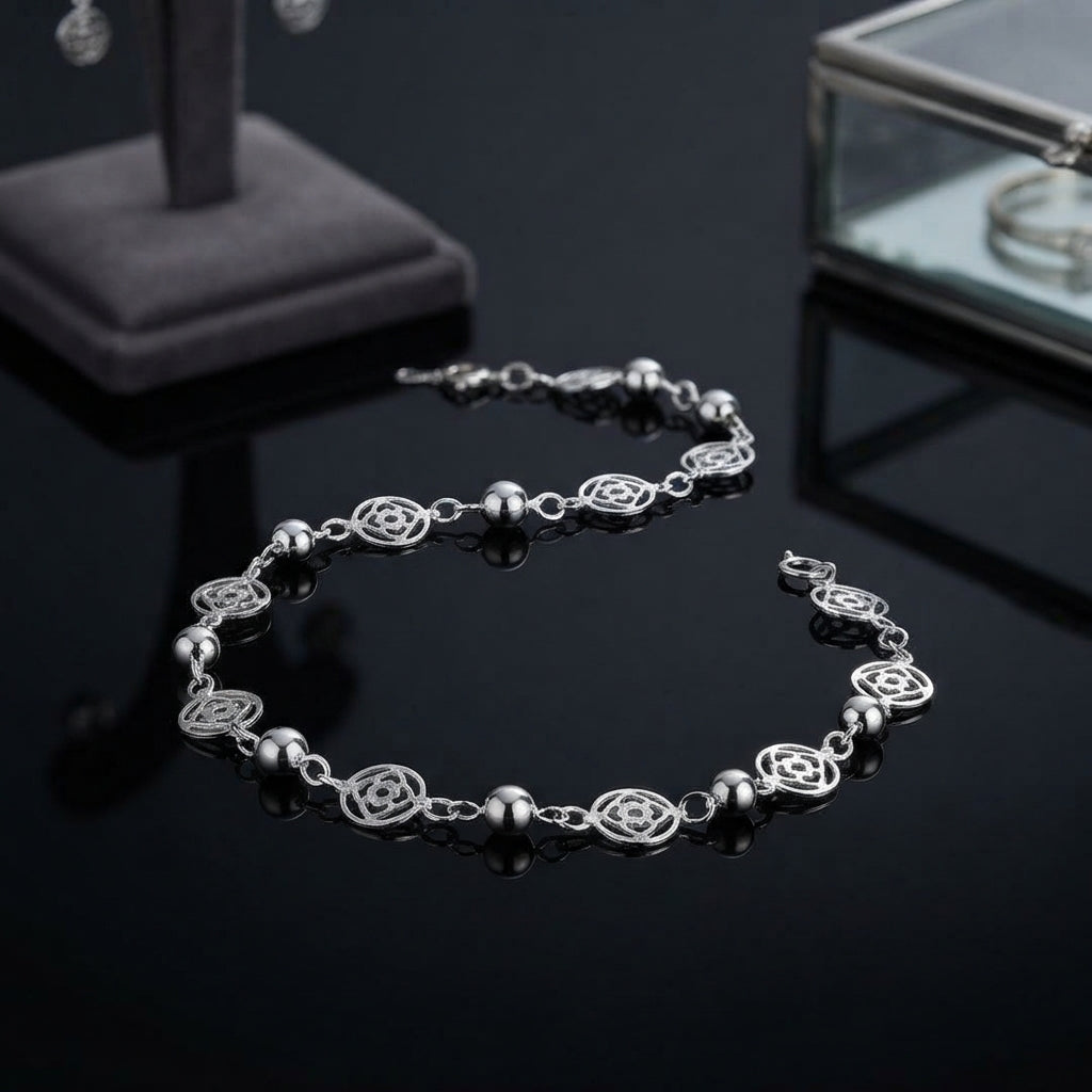 Silver Filigree Flower Bead Station Chain Anklet on black mirror surface with jewellery display stand and glass box in background showing fine silver cable chain with alternating round smooth silver ball beads and open oval filigree flower rose motif charms in station-link design with lobster clasp and mirror reflection below