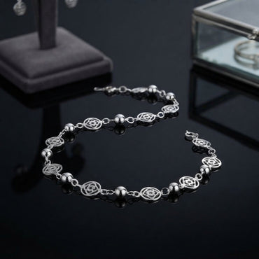 Silver Filigree Flower Bead Station Chain Anklet on black mirror surface with jewellery display stand and glass box in background showing fine silver cable chain with alternating round smooth silver ball beads and open oval filigree flower rose motif charms in station-link design with lobster clasp and mirror reflection below