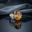 Silver Imperial Citrine Oval CZ Pavé Halo Flower Shoulder Cocktail Ring — front 3/4 elevated view: oversized golden citrine oval with CZ pavé halo, 5-petal CZ flower cluster shoulders, slim pavé band, on dark mirror surface with grey silk fabric backdrop