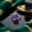 Silver-Tone Large Round Amethyst Purple CZ Solitaire Bypass Pavé Band Statement Ring on black reflective surface with emerald green gold-embroidered fabric and navy blue gold jewelry box backdrop