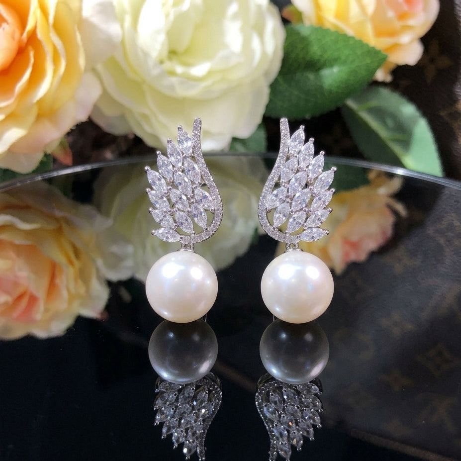 Silver-Tone Marquise CZ Angel Wing Feather Cluster & Large Pearl Drop Earrings on black mirror surface with orange roses and green leaves backdrop