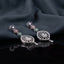 Silver Multi-Color CZ Circle Chain and Open Oval Filigree Pear Drop Earrings — rhodium silver-tone bridal earrings with 3-tier sapphire blue ruby red and emerald green baguette CZ circle disc chain open black rhodium oval honeycomb filigree pendant with large pear white CZ center and pear CZ tip drop displayed on dark black mirror surface with charcoal satin backdrop