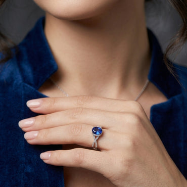 Silver-Tone Oval Blue Sapphire CZ Halo & Infinity Crossover Pavé Band Ring worn on right hand of female model in royal blue velvet blazer with grey studio backdrop