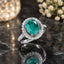Silver-Tone Oval Emerald CZ Scalloped Halo & Pavé Shoulder Cocktail Ring on dark green marble surface with bokeh gold light orb and white baby's breath flowers backdrop