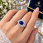 Silver-Tone Oval Royal Sapphire Blue CZ Micro-Pavé Inner Halo & Baguette Sunburst Outer Halo Adjustable Cocktail Ring worn on model hand with bridal lace outfit, white flowers and Destiny navy jewelry box in background
