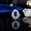 Silver-Tone Oval Sapphire Blue CZ Ballerina Baguette Sunburst & Pavé Double Halo Bridal Statement Ring on black mirror surface with white crystal quartz cluster and royal blue velvet backdrop